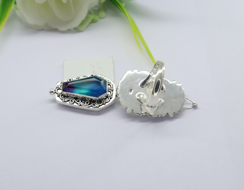 K&ouml;nnte beinhalten: Ein silberner Ring mit einem gro&szlig;en, facettierten, blauen und violetten Edelstein, der in einem detaillierten, verzierten Design gefasst ist. Der Ring wird von der Seite und von hinten gezeigt.