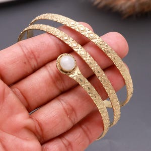 May include: A gold-toned metal wrap bracelet featuring a repeating diamond-shaped textured pattern along the band. The terminal end of the bracelet is finished with a single, smooth, round white stone set within a twisted rope-style metal border.
