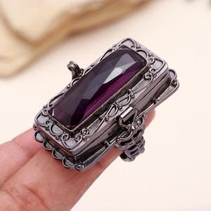 May include: A silver ring with a hinged box top and a large, rectangular, purple gemstone. The box is decorated with intricate scrollwork.