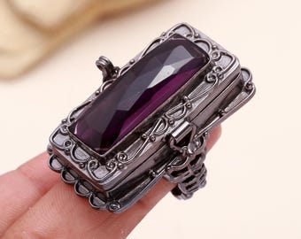 May include: A silver ring with a hinged box top and a large, rectangular, purple gemstone. The box is decorated with intricate scrollwork.