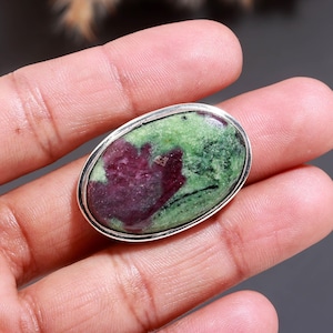 May include: An oval-shaped ring featuring a polished cabochon stone with a mottled pattern of deep magenta and forest green. The gemstone is set within a smooth, polished silver-toned metal bezel that follows the rounded contour of the stone.