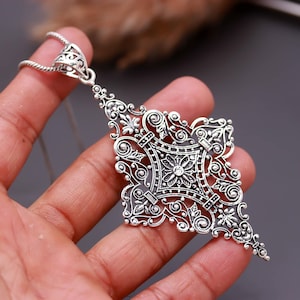 May include: A silver-toned pendant necklace featuring an intricate, diamond-shaped filigree design with ornate scrollwork, floral motifs, and a small central clear stone. The pendant hangs from a thin, textured silver-toned chain.