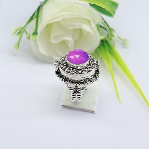 Op de afbeelding: Een zilveren ring met een scharnierend compartiment en een paarse edelsteen. De ring is versierd met ingewikkelde bloemmotieven.