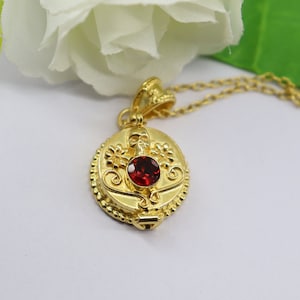 May include: A gold pendant necklace with a round locket featuring a red gemstone and a skull design. The locket has a detailed floral pattern and a beaded edge.