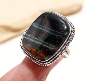 May include: A silver ring with a large, square-shaped, dark green and brown tiger's eye gemstone. The ring has a decorative, braided silver band.