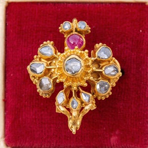 May include: A gold ring with a red gemstone in the center and a cluster of smaller, clear gemstones around it. The ring is set in a floral design.