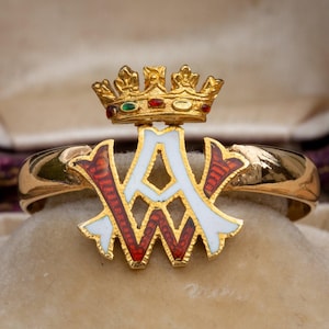 May include: A gold ring featuring a white enamel monogram and a crown with red, green, and gold enamel details.