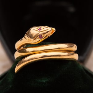 May include: A gold-colored snake ring with ruby-colored eyes. The snake's body coils around, creating a spiral design. The ring is displayed on a dark green velvet surface, set against a black background.