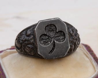 May include: A silver ring with a detailed design featuring a shamrock. The ring is on a pink and white ring box.