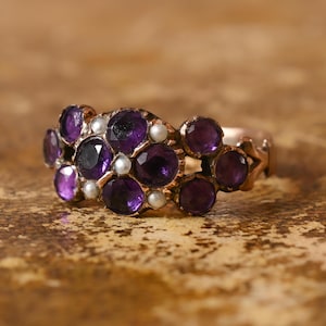 Puede incluir: Un anillo vintage con un grupo de piedras preciosas de color púrpura intenso y pequeñas perlas blancas. El anillo tiene una banda dorada y un diseño floral en los lados. Las piedras preciosas están dispuestas en un patrón floral.