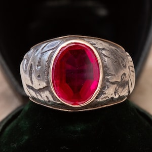 May include: A silver ring featuring a large, oval, bright red gemstone. The ring's band is textured with a leaf pattern. The gemstone is set in a gold-coloured bezel. The ring is presented in a black jewellery box.