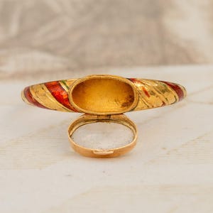 May include: A gold-toned hinged bangle bracelet with red and gold enamel stripes. The bracelet is open, revealing a hollow interior. A small, matching gold-toned locket is open below the bracelet.