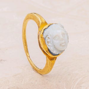 May include: A gold ring with a white cameo featuring a carved face. The cameo is set in a gold bezel.