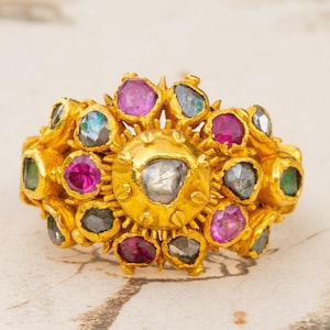 Puede incluir: Un anillo de oro con un gran diamante de talla rosa, facetado, en el centro, rodeado de diamantes, rubíes y esmeraldas más pequeños.