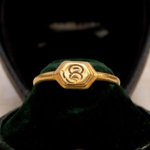 May include: A gold ring with a hexagonal face featuring an engraved design, set against a dark green velvet cushion inside a black jewelry box. The ring's band is thin and polished.