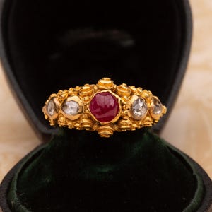 May include: A gold ring with a central ruby and surrounding clear gemstones. The ring is set in a detailed, ornate design. The ring is displayed in a black jewelry box with a green velvet interior.