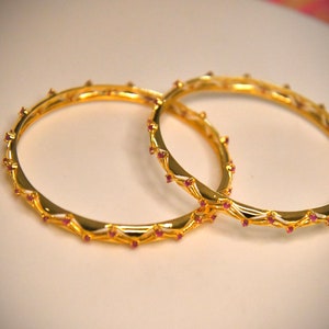 Könnte beinhalten: Zwei goldene Armreifen mit einem dekorativen Design und kleinen roten Edelsteinen.