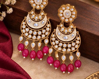 May include: A pair of gold-toned chandelier earrings featuring a floral stud, a crescent-shaped drop with white enamel detailing, inset clear stones, and dangling teardrop-shaped fuchsia beads suspended from the bottom edge of the metal frame.