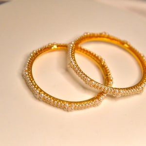 Pode incluir: Dois braceletes de ouro com uma fila de pérolas brancas ao redor da borda.