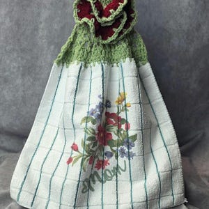 May include: A handmade kitchen towel with a green crocheted top and a burgundy flower accent. The white towel features a floral design with the word "mom" and blue and green stripes.