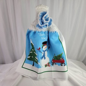 May include: A white kitchen towel with a blue crocheted top and a snowman design. The towel features the text "Tis' the Season to be Jolly!" and a snowman pulling a red wagon with a Christmas tree.
