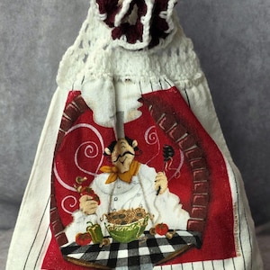 May include: White kitchen towel with a crochet top and a burgundy flower. The towel features a chef illustration with a bowl of spaghetti, tomatoes, and peppers on a checkered tablecloth.