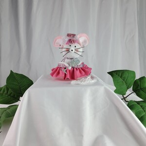 May include: A white sock mouse wearing a pink dress with a sparkly silver heart and a pink flower crown. The mouse is sitting on a white surface.