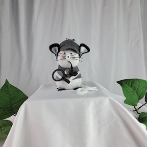 May include: A gray and white sock mouse wearing a detective hat and holding a magnifying glass. The mouse is sitting on a white surface with a white string.