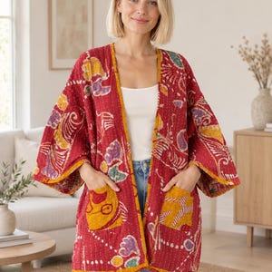 Könnte beinhalten: Eine rote, vorne offene Jacke mit einem mehrfarbigen Batikmuster in Gelb, Lila und Weiß. Die Jacke hat weite Dreiviertelärmel, einen gelben Saum an den Kanten und zwei große aufgesetzte Taschen auf der Vorderseite.