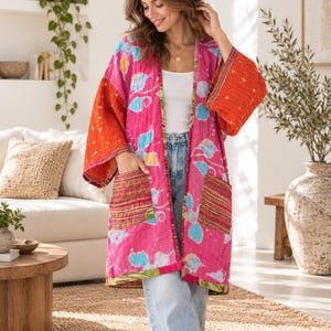 May include: A long-sleeved open-front jacket featuring a vibrant pink base with a multi-coloured floral pattern, contrasting orange sleeves, and two front patch pockets with horizontal stripes. The garment has a quilted texture and loose fit.