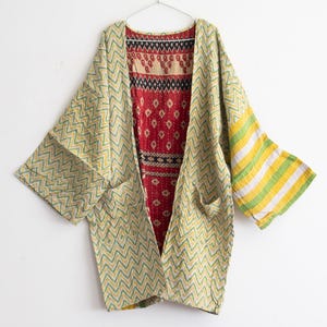 May include: A reversible open-front jacket featuring a yellow and green chevron pattern on one side and a red interior with intricate black and white geometric and paisley patterns. The garment includes two front pockets and wide sleeves.
