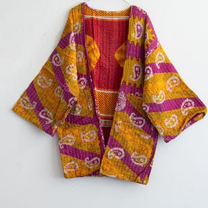May include: A colorful, open-front kimono-style jacket. The jacket features a vibrant mix of yellow, purple, and red fabrics with a paisley pattern. The jacket is hanging on a metal hanger against a white background.
