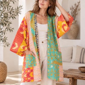 Può includere: Una giacca kimono a maniche lunghe con apertura frontale, caratterizzata da un design patchwork con motivi vivaci nei toni del rosso, giallo e verde acqua. Il capo presenta due tasche applicate sul davanti e cuciture visibili su tutto il tessuto.