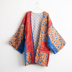 May include: A vibrant, open-front kimono jacket. It has a patchwork design with red, blue, and gold floral patterns. The sleeves are wide, and the jacket is hanging on a silver hanger against a white background.