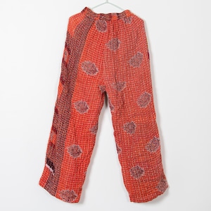 May include: Loose-fitting, orange pants with a patchwork design. The pants feature a red and brown patterned panel on the left side and a repeating floral pattern across the orange fabric. The pants are hanging on a white hanger.