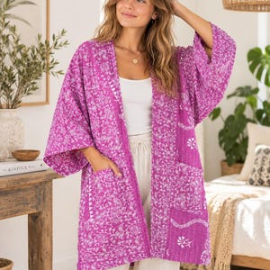 Pode incluir: Um casaco tipo quimono de cor magenta vibrante com frente aberta, apresentando um padrão intrincado de flores e vinhas brancas por toda a peça. Esta vestimenta leve inclui mangas largas de três quartos e dois bolsos frontais grandes.