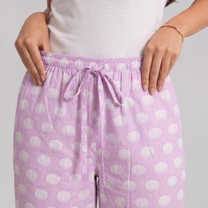 May include: Light purple lounge pants featuring an all-over white seashell pattern. The pants have an elasticized waistband with a matching fabric drawstring tie centered at the front, providing a secure and adjustable fit for the wearer.