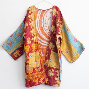 May include: A colorful, open-front kimono-style jacket with a patchwork design. The jacket features a mix of red, yellow, orange, and blue floral patterns. It has two front pockets and is hanging on a silver hanger.