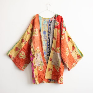 May include: A long-sleeved open-front jacket featuring a patchwork design with orange, yellow, and pink floral patterns. The garment includes two front pockets and visible stitching throughout, hanging on a wire hanger against a plain background.