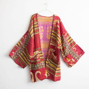 May include: A red, yellow, and pink patterned kimono-style jacket with wide sleeves. The jacket is made of quilted fabric and features a floral and geometric design. It is hanging on a white hanger against a white background.