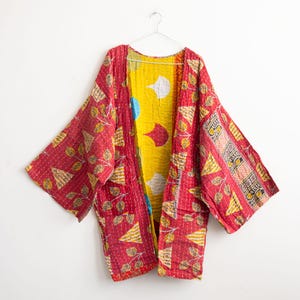 May include: A red and yellow patterned kimono jacket hanging on a white hanger. The jacket has a patchwork design with red, yellow, and white patterns. The interior lining is bright yellow with white and red shapes.