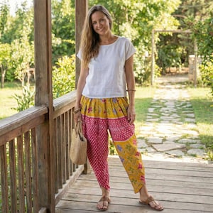 May include: A woman wearing a white short-sleeve top and colorful patterned pants. The pants feature a combination of pink, yellow, and floral designs. She is holding a woven tote bag and wearing sandals. The background includes a wooden porch and lush greenery.