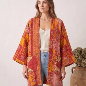 Puede incluir: Una chaqueta acolchada estilo kimono con un diseño de patchwork en tonos rojos, naranjas y amarillos. La prenda tiene mangas anchas, frente abierto y dos bolsillos de parche delanteros, decorados con patrones florales y geométricos.