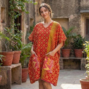 May include: A red and gold patterned kaftan dress with a V-neck and short sleeves. The dress features two front pockets with a contrasting pink and yellow paisley design. The dress is knee-length and made of a lightweight fabric.