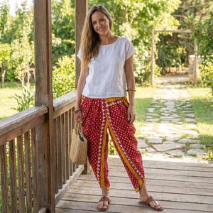 Op de afbeelding: Een vrouw draagt een witte top met korte mouwen en een rode broek met een geel en wit patroon. Ze houdt een strooien tas vast en draagt sandalen. De achtergrond omvat een houten veranda en groen.