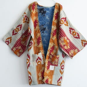 May include: A long-sleeved open-front jacket featuring a multi-colored ikat pattern in shades of orange, red, and cream. The interior lining is a contrasting blue fabric with a dark circular print. Two patch pockets are on the front.