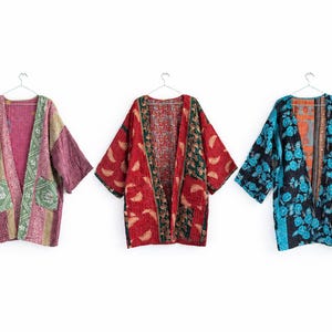 May include: Three open-front jackets with wide sleeves, each featuring distinct patchwork patterns. The left jacket is pink and green, the center is red and dark green, and the right is blue and black with floral motifs, all hanging on hangers.