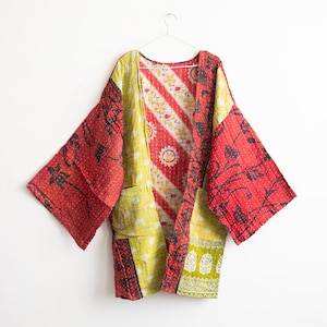 Pode incluir: Casaco estilo quimono acolchoado e colorido com design patchwork, pendurado num cabide branco. O casaco apresenta painéis vermelhos, amarelos e brancos com estampados florais e geométricos. Tem mangas largas e dois bolsos frontais.