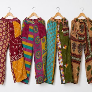 Puede incluir: Cuatro pares de pantalones patchwork coloridos cuelgan de perchas de madera sobre un fondo blanco. Los pantalones presentan varios patrones, incluyendo diseños florales, geométricos y a rayas, en tonos rojos, amarillos, verdes, azules y marrones. Los pantalones parecen estar hechos de una tela suave y ligera.