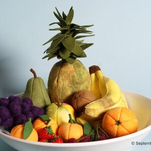 Op de afbeelding: Een witte keramische kom gevuld met verschillende viltfruit, waaronder een ananas, een peer, druiven, bananen, sinaasappels en appels. Het fruit is in een decoratief patroon gerangschikt.